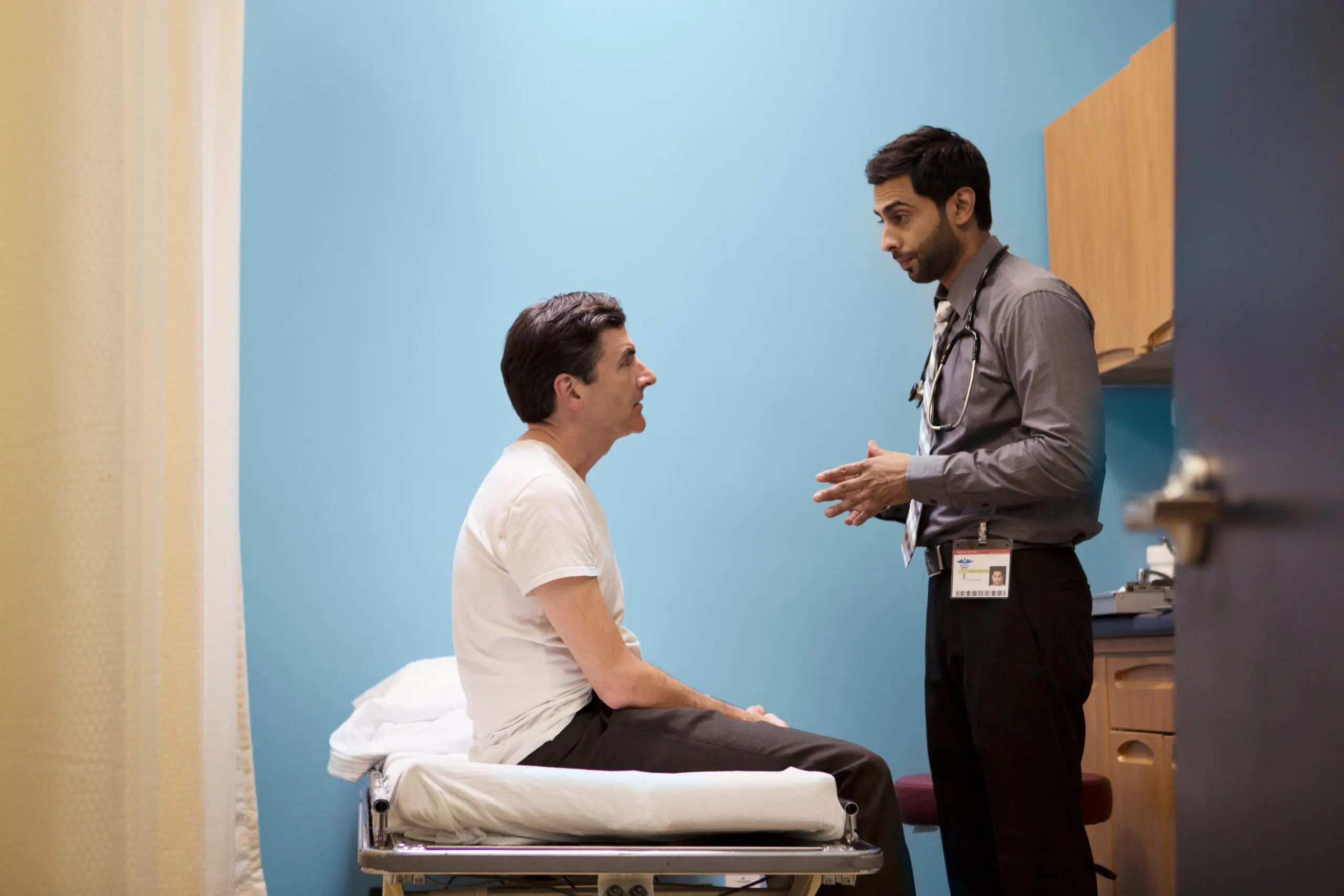 A young male doctor speaks to a patient sitting on a bed.