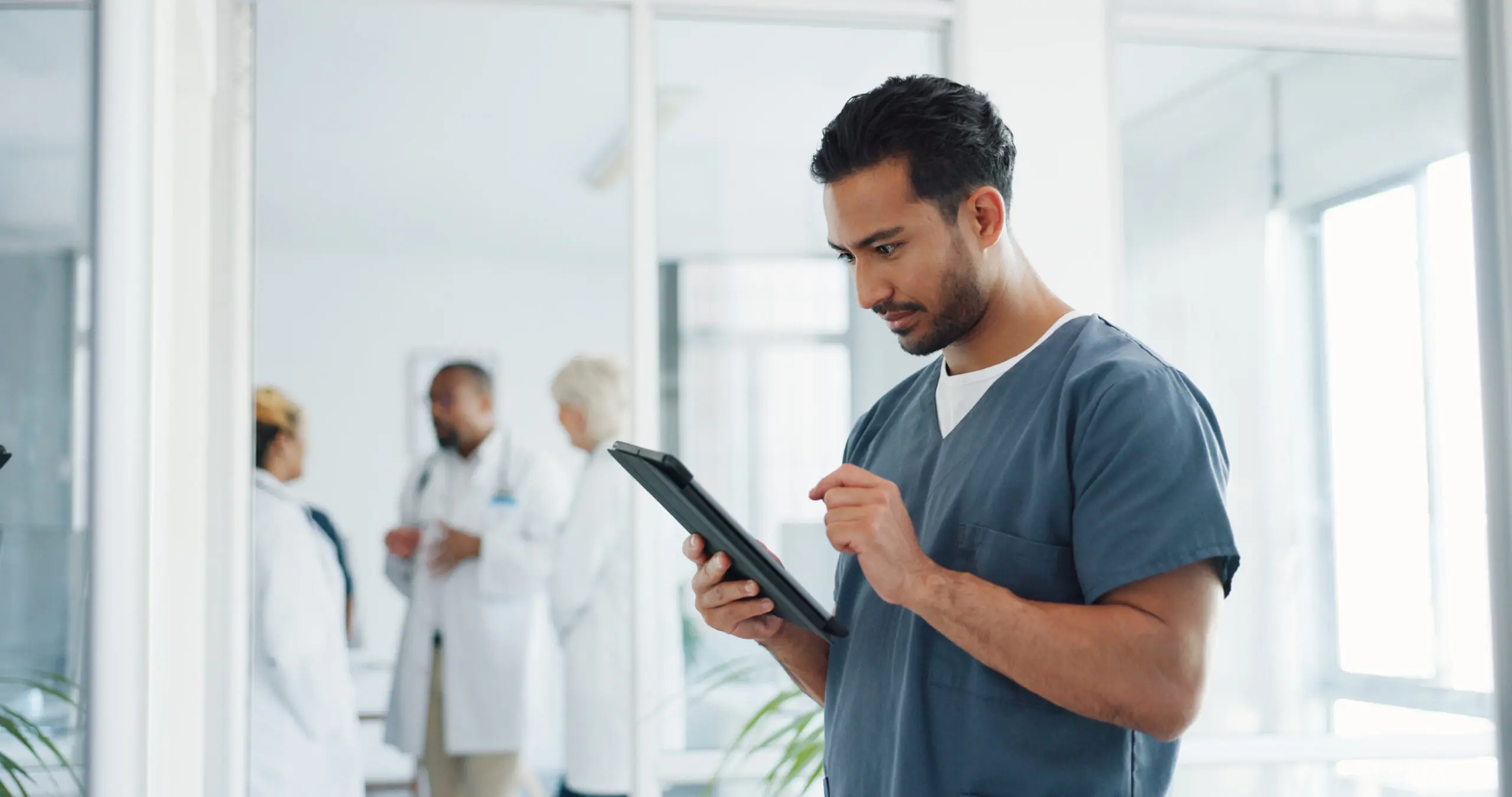A doctor interacts with a tablet, as a group of other doctors speak in the room behind.